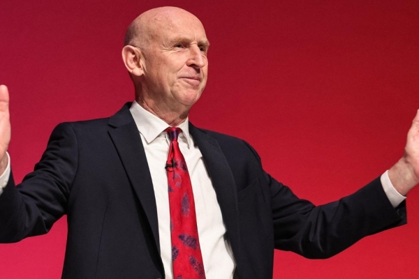 Chancellor John Healey - a white man wearing a dark suit - stood against a red background with his arms open wide.